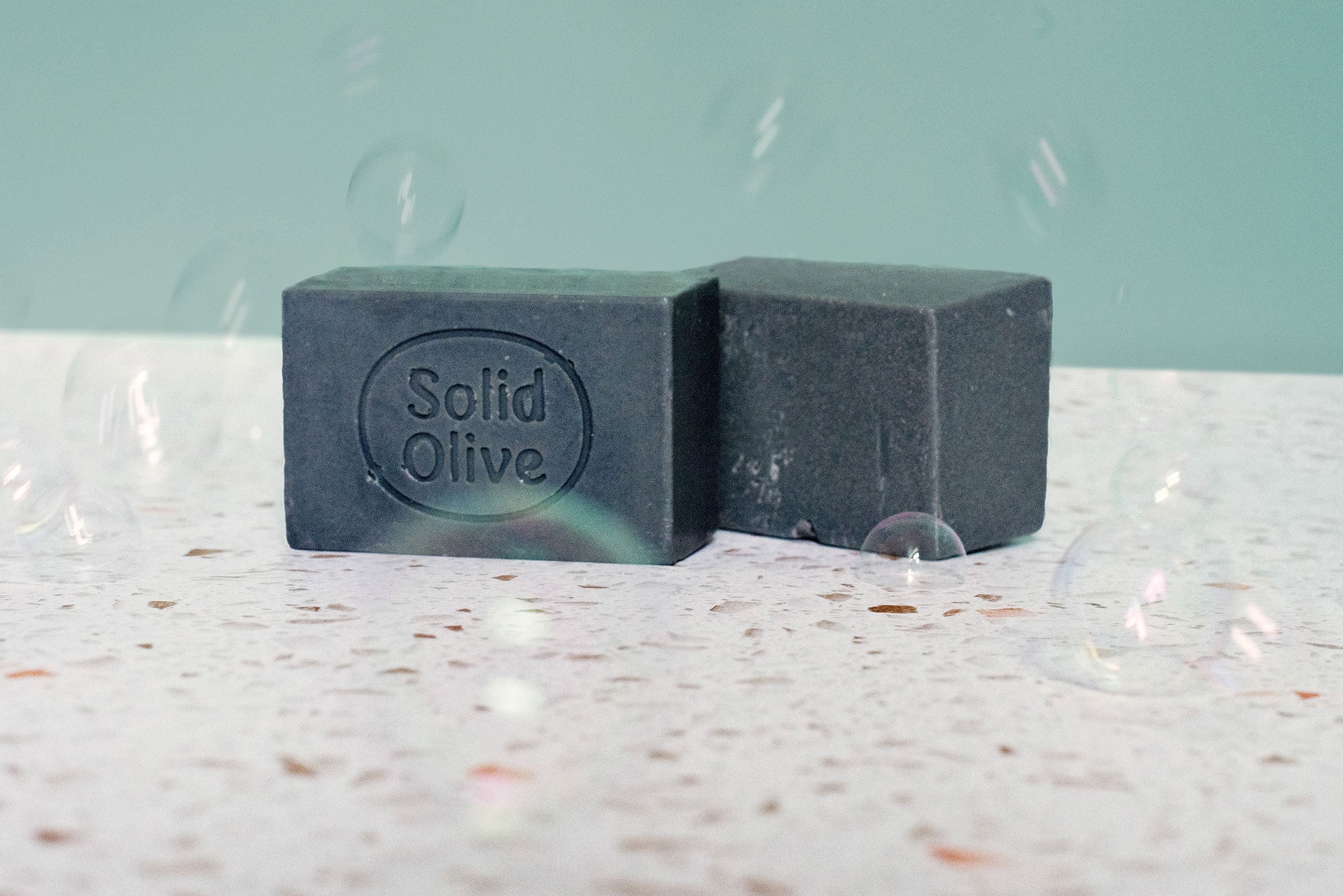 Two bars of Castile soap made from activated charcoal sit on a terrazzo countertop. Bubbles float around them.