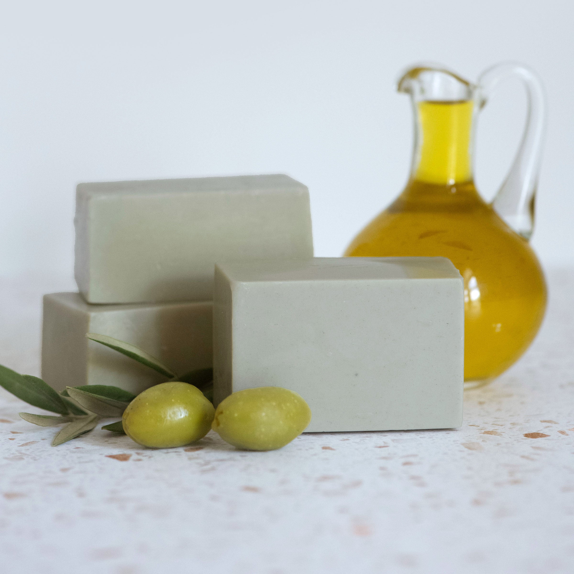 Three bars of green, Castile soap on a countertop. Olives and olive oil are in the background.