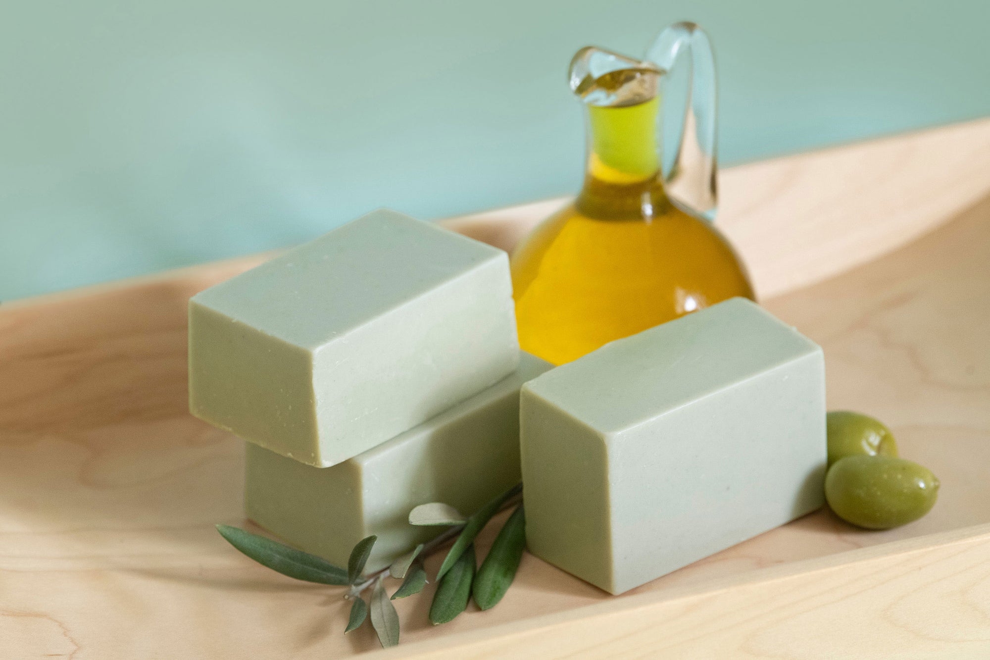 Three bars of Castile soap sit in a light wooden tray. Olives and olive oil are next to them.