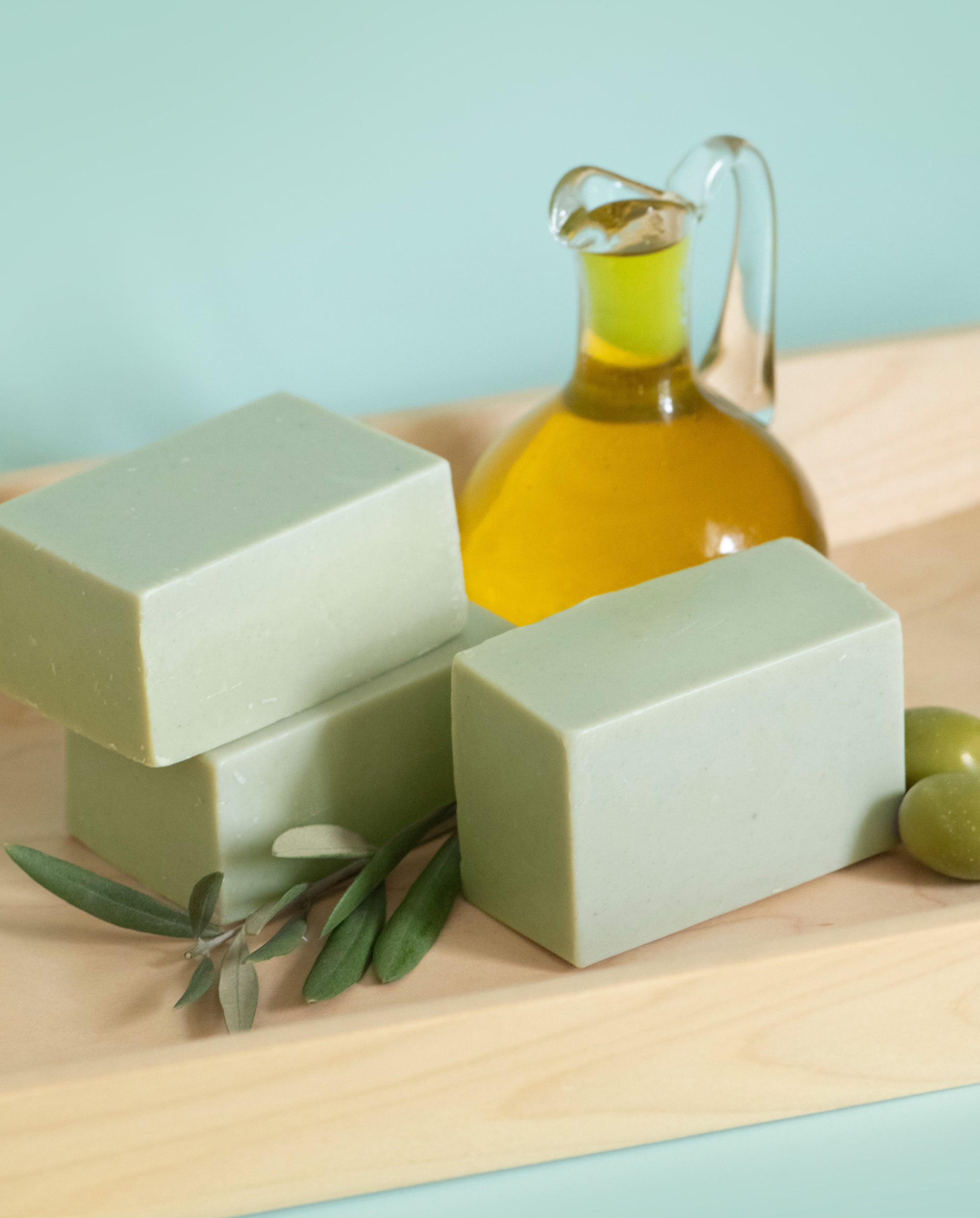 Three bars of green soap with a bottle of olive oil on a light wooden surface.