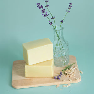 Two bars of baby soap sit on a wood tray. Oats and lavender lay next to it.