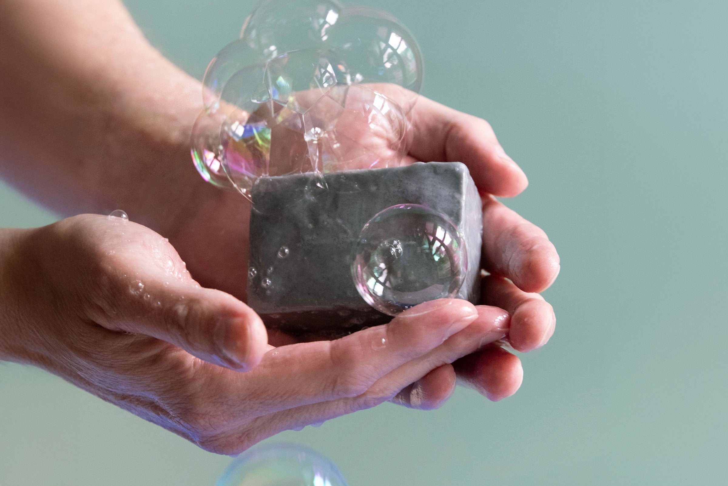 Hands hold a bar of Castile soap made from activated charcoal. Bubbles float around it.