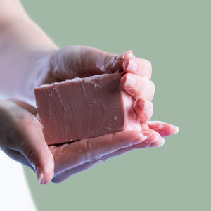 Hands holding a sudsy pink bar of Castile soap against a green background.