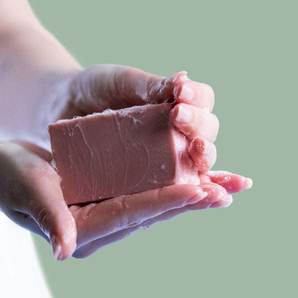 Hands holding a sudsy pink bar of Castile soap against a green background.