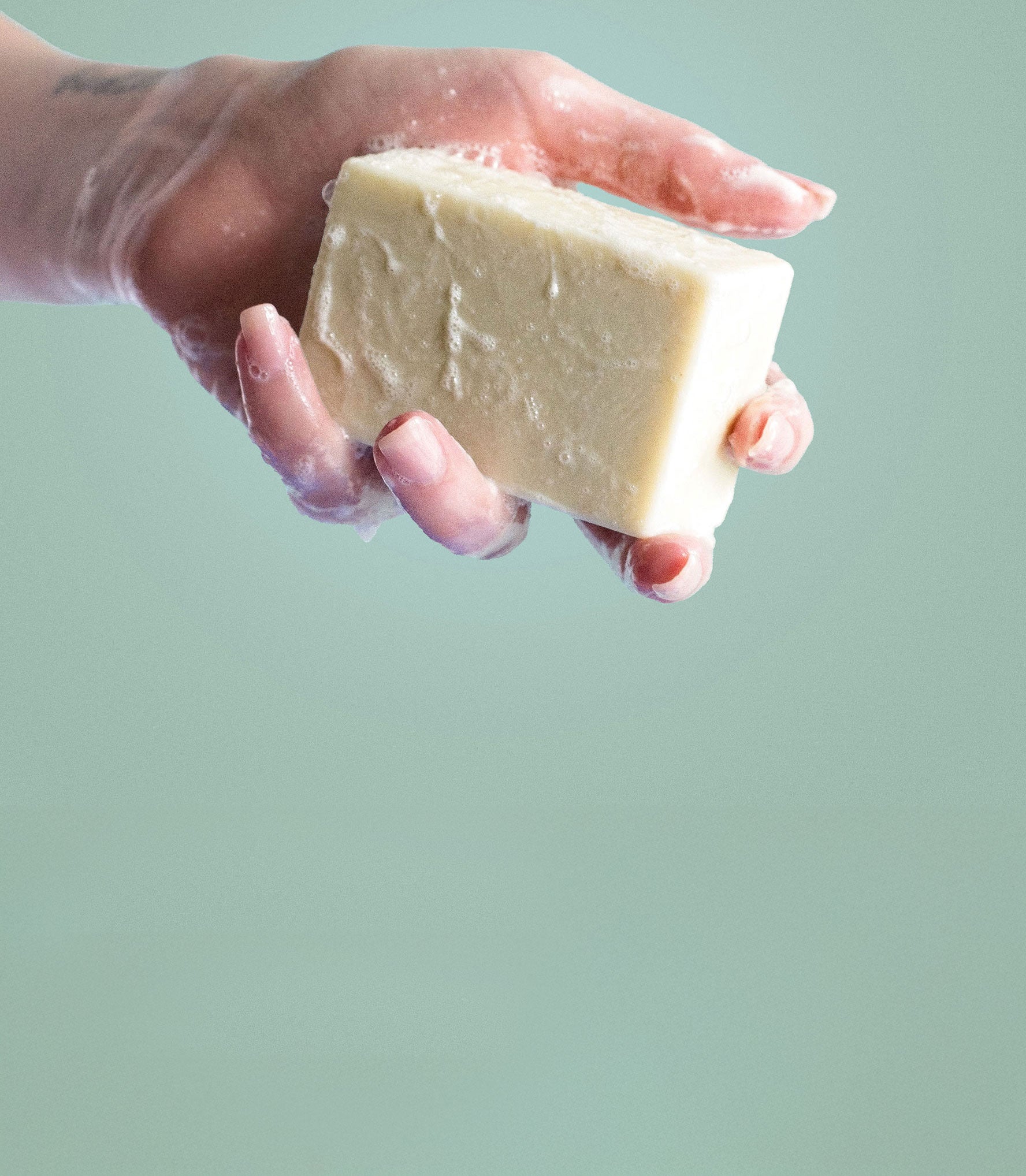 A hand holding a bar of unscented castile soap against a green background.
