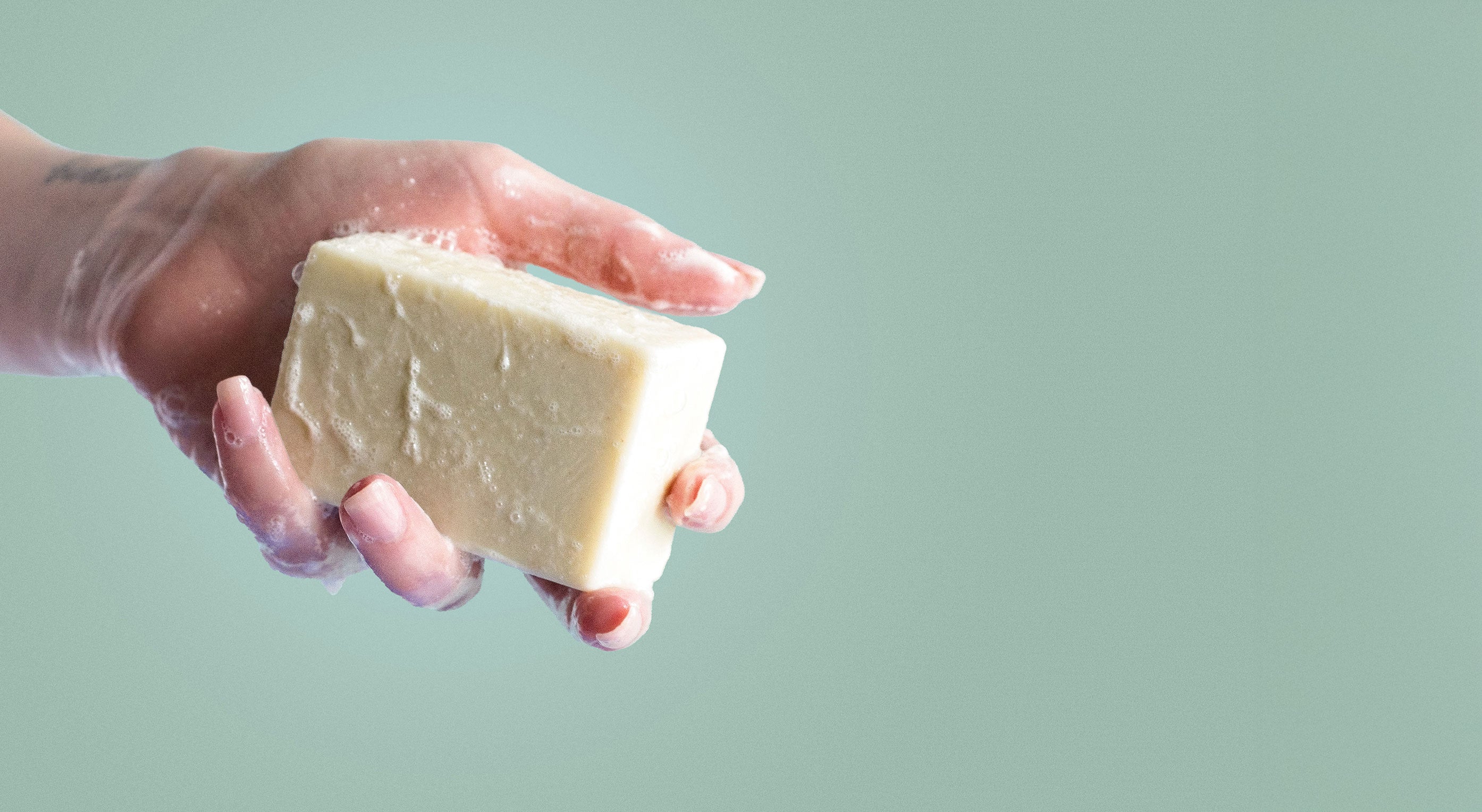 A hand holds a sudsy bar of unscented castile soap against a green background.