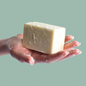 A hand holds up a bar of unscented castile soap. Both have soap suds on them.