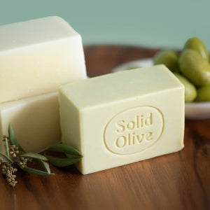 Bars of unscented castile soap sits on a wooden tray with olives and olive leaves next to it. One has a Solid Olive logo on it.