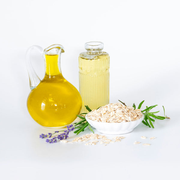 Olive and castor oil in glass vases sit on a white background next to oats in a white bowl. Lavender flowers and leaves poke through.