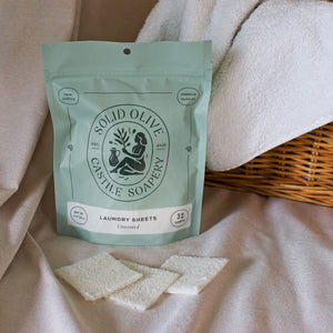A bag of laundry sheets sits in front of a wicker basket on a linen tablecloth. Three laundry sheets are laid in front of it.