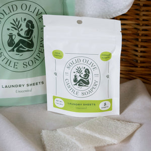 A bag of unscented laundry sheets with a Solid Olive logo on it sits on a linen cloth. A towel and basket can be seen in the background.