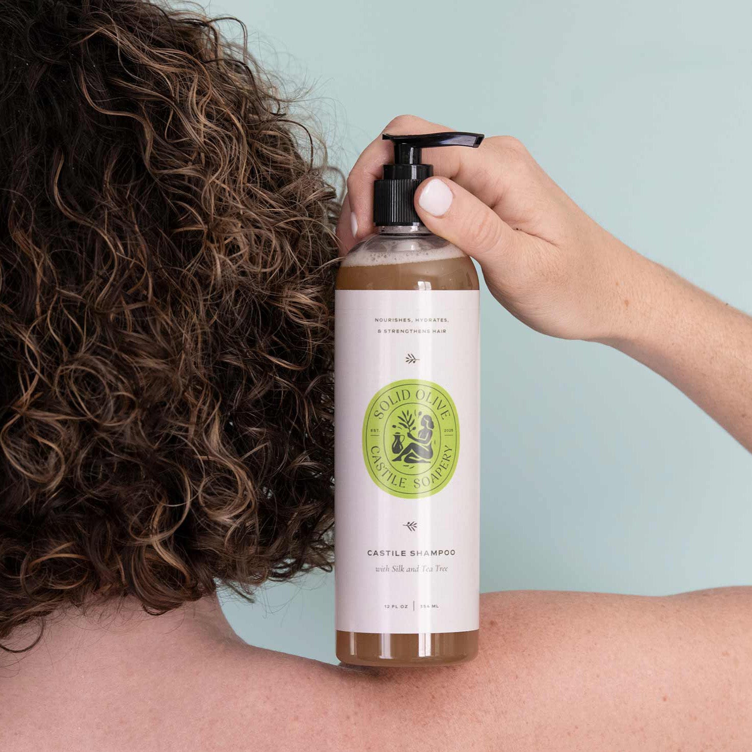Person holding a bottle of Castile shampoo with a visible label against a light blue background